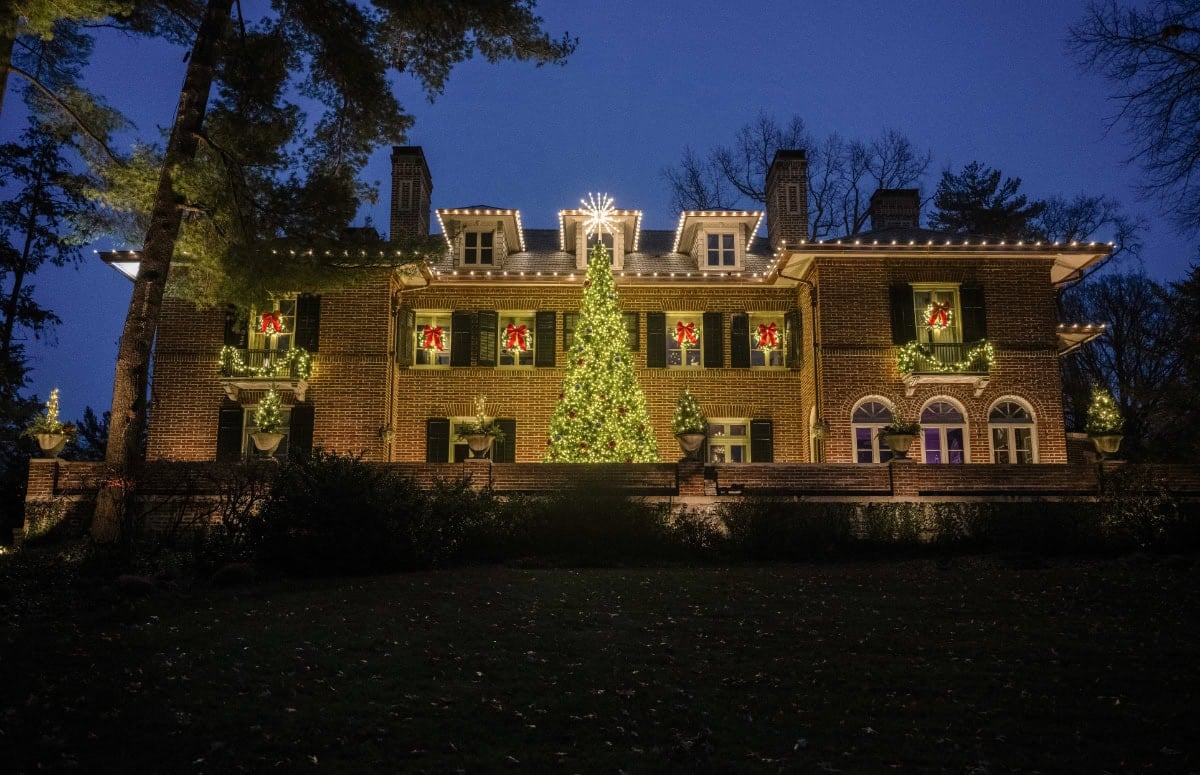 Large brick house decorated with Christmas lights, wreaths, and a large lit tree in the center at night. - Light Up Columbus