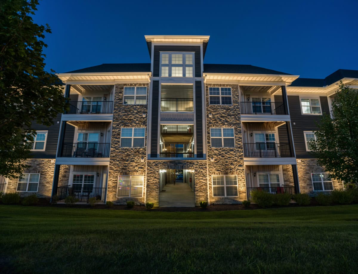 A modern three-story apartment building with stone and siding exterior, illuminated at night, viewed from a grassy lawn. - Light Up Columbus