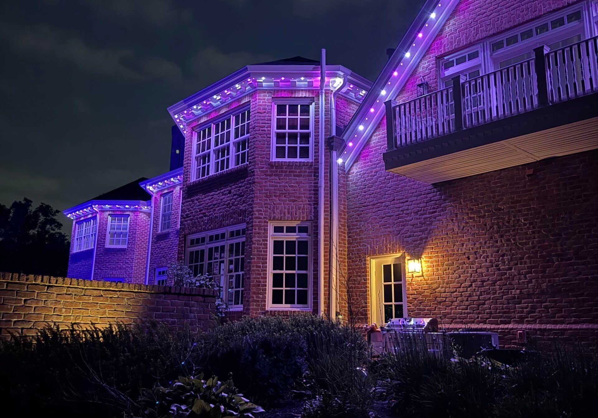 A brick house at night with purple and white LED lights outlining the roof, a lit wall lamp near the back door, and cloudy sky overhead. - Light Up Columbus